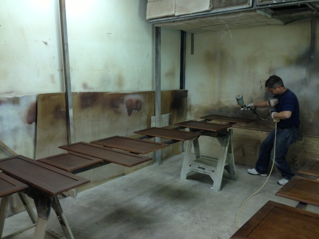 A person spray-paints wooden cabinet doors arranged on saw horses in a workshop.