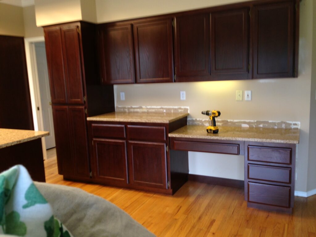 Kitchen scene with dark wood cabinets, beige countertops, and a power drill on a lower counter. Walls have unpainted patches.