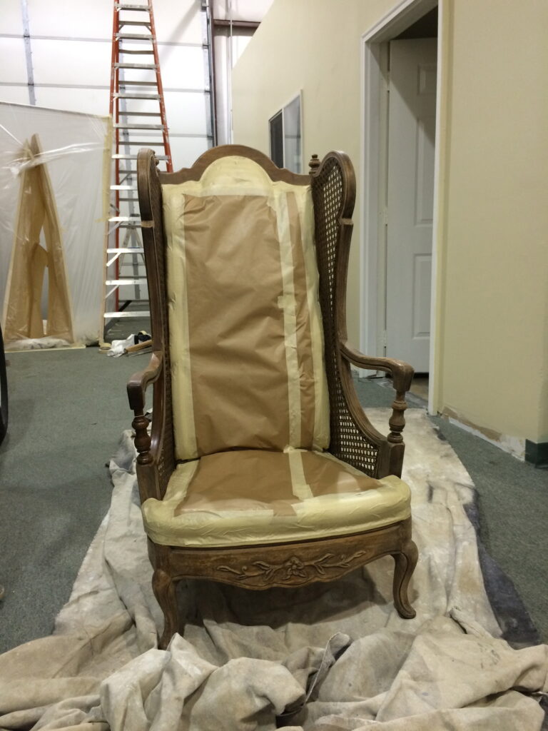 A vintage wooden chair with paper covering is situated indoors, with a ladder and door in the background.