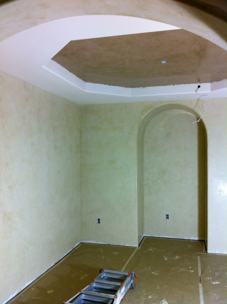A room under construction with beige walls, a recessed ceiling, an alcove, and electrical wiring exposed. Ladder on floor.