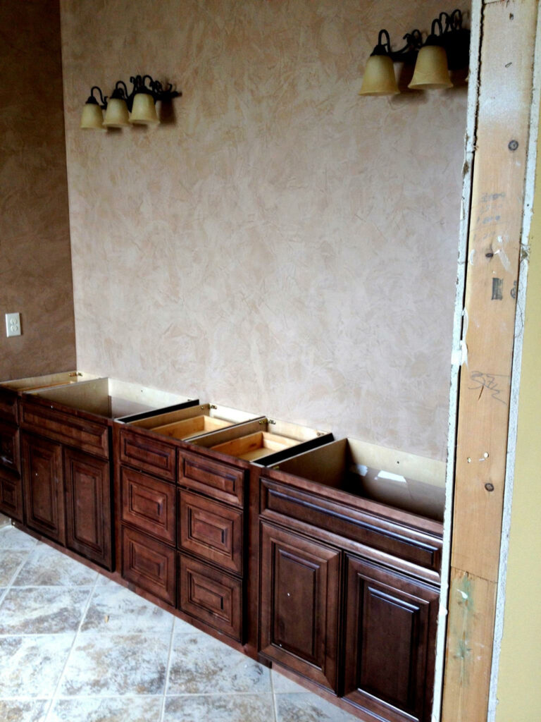 Unfinished bathroom renovation with wooden cabinets installed and light fixtures on a textured wall above tiled flooring.
